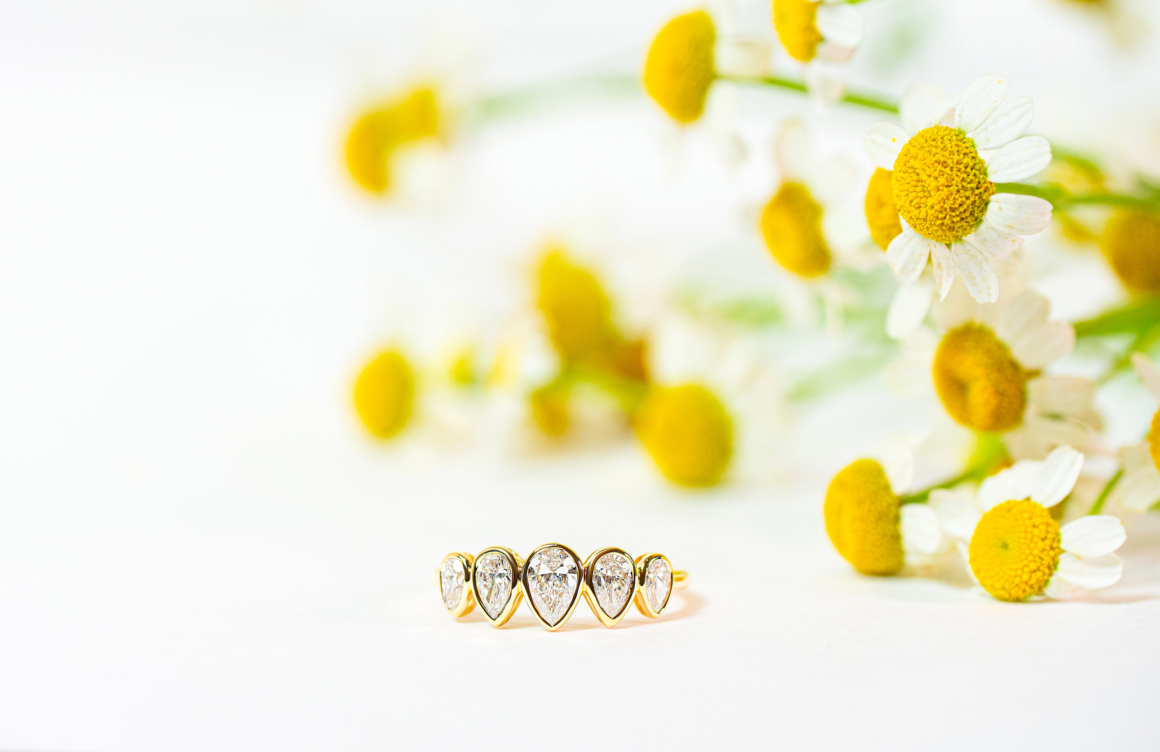 Gold ring with pear-shaped diamonds on a white background with yellow flowers.