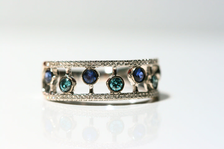 Band style ring. Bezel set sapphires float between thin bands of white diamonds. The sapphires are alternating dark blue and teal. The open spaces between the stones create interesting shapes on the finger. This design is only on the front half of the ring, the back half is solid white gold. Local SF jewelry, San francisco, Luxury, hand made, Bay area, gift for her, cocktail ring.