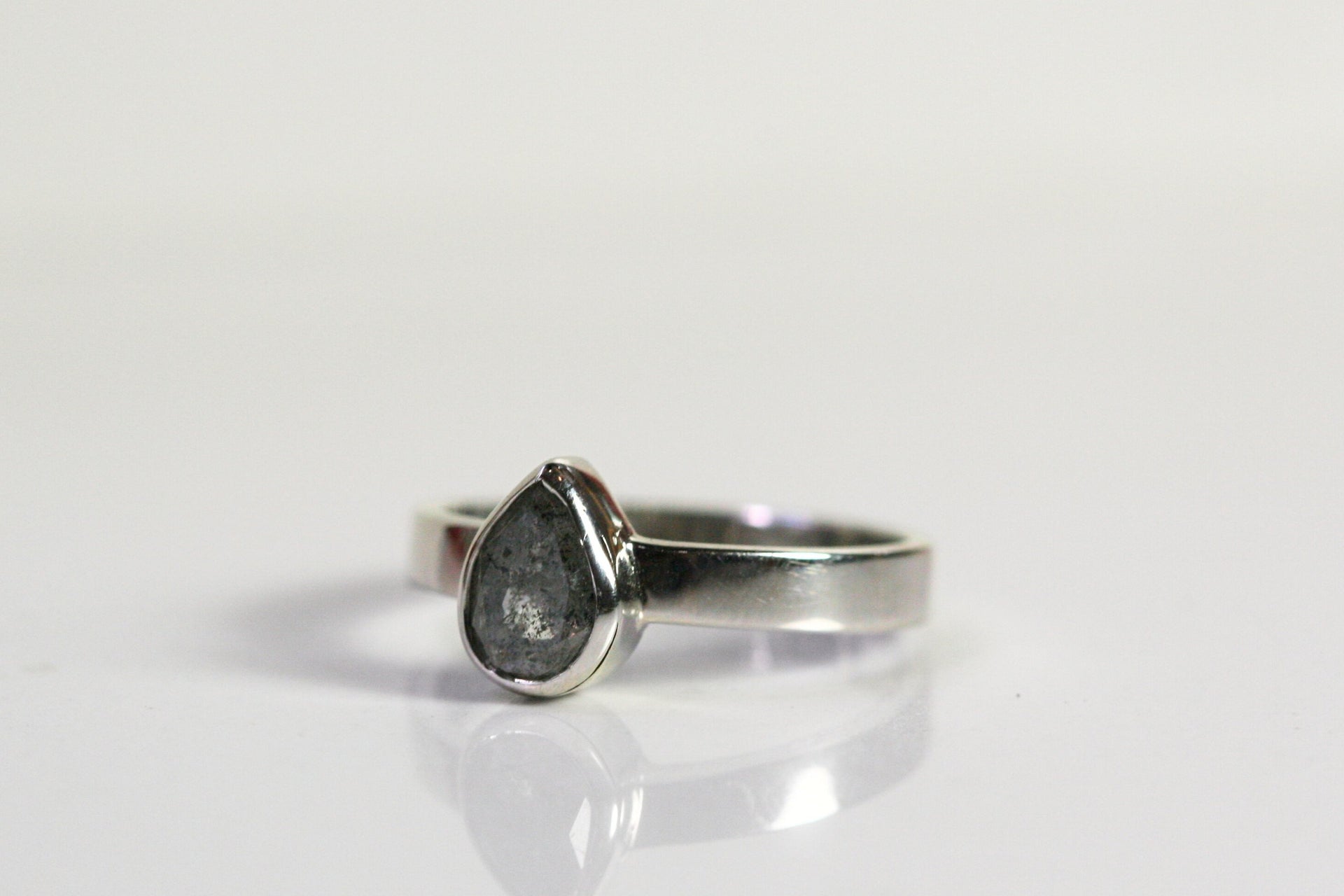 A pear shaped salt and pepper diamond is bezel set in white gold. The band is thick. The stone has interesting inclusions.