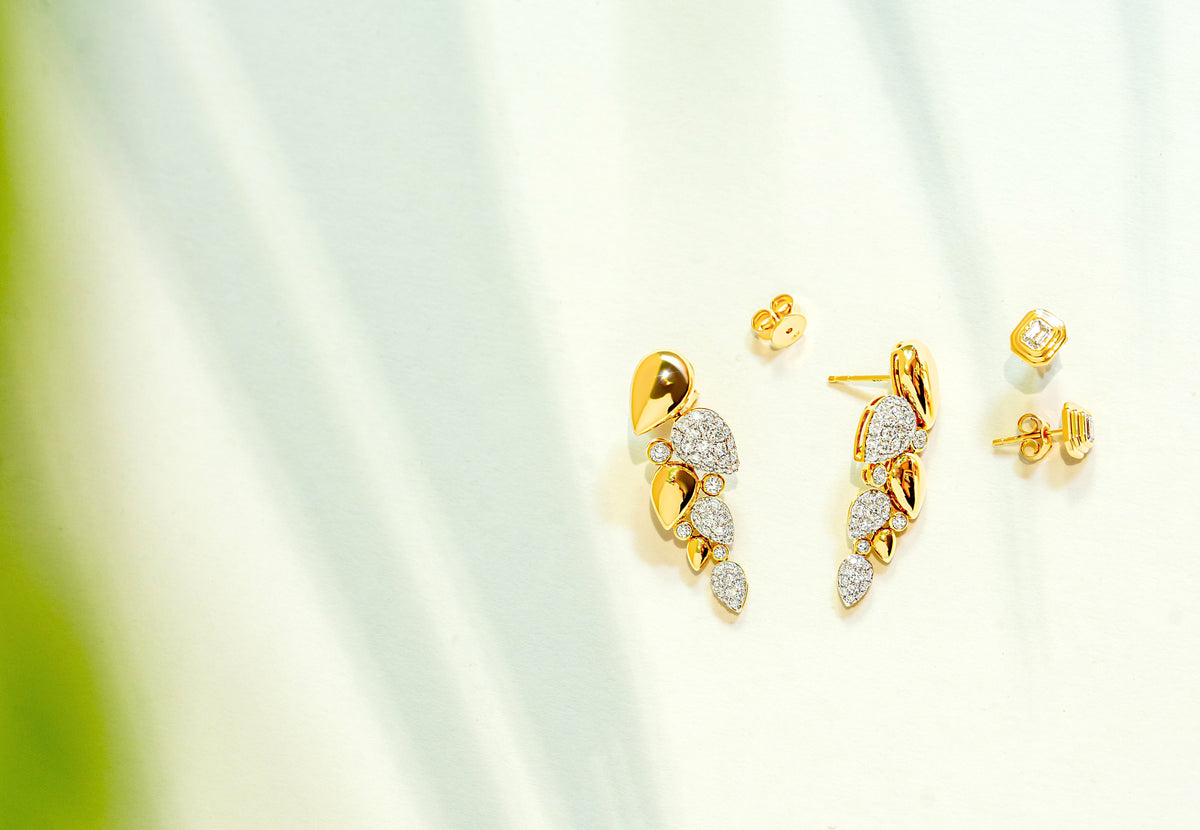 Gold and diamond leaf-shaped drop stud earrings displayed on a soft white background with subtle green leaf shadows.