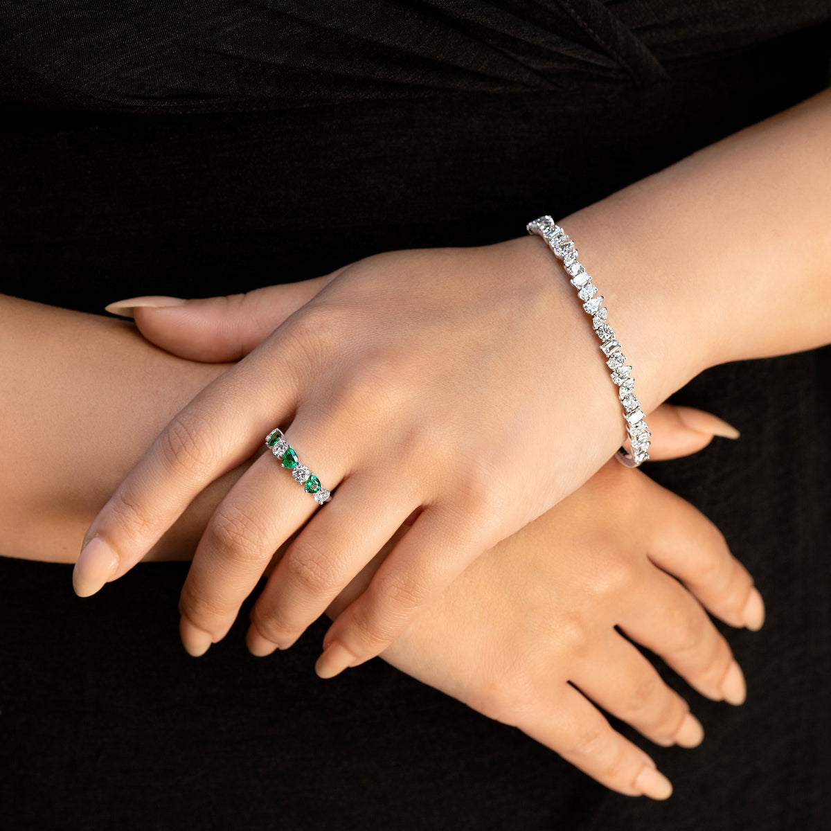 A close-up of a person's hands wearing a silver mixed-cut diamond tennis bracelet on one wrist and a matching silver ring with green emerald-cut stones on the finger, set against a black background.