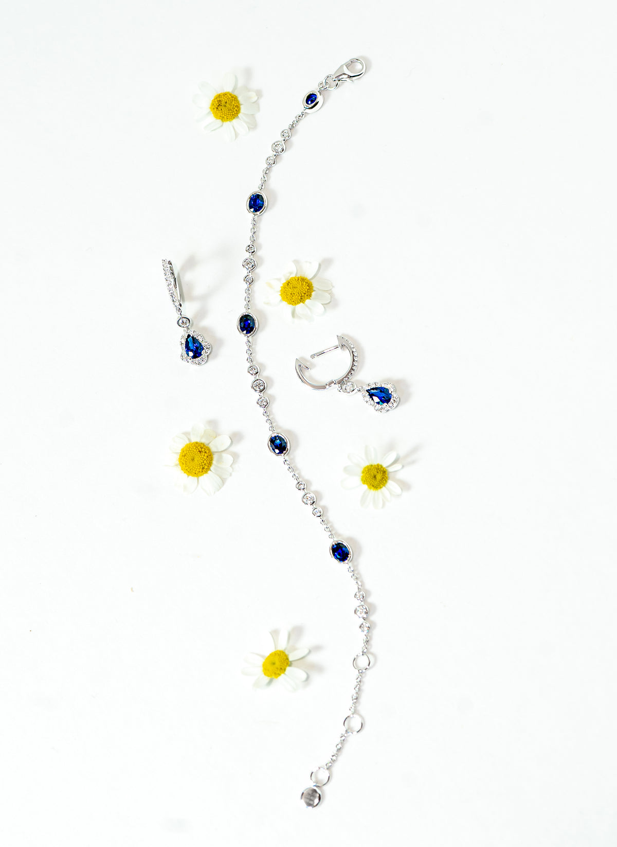 A silver-toned jewelry set featuring a bracelet, matching earrings, decorated with small blue sapphires and white diamonds, styled with daisy flowers on a white background.