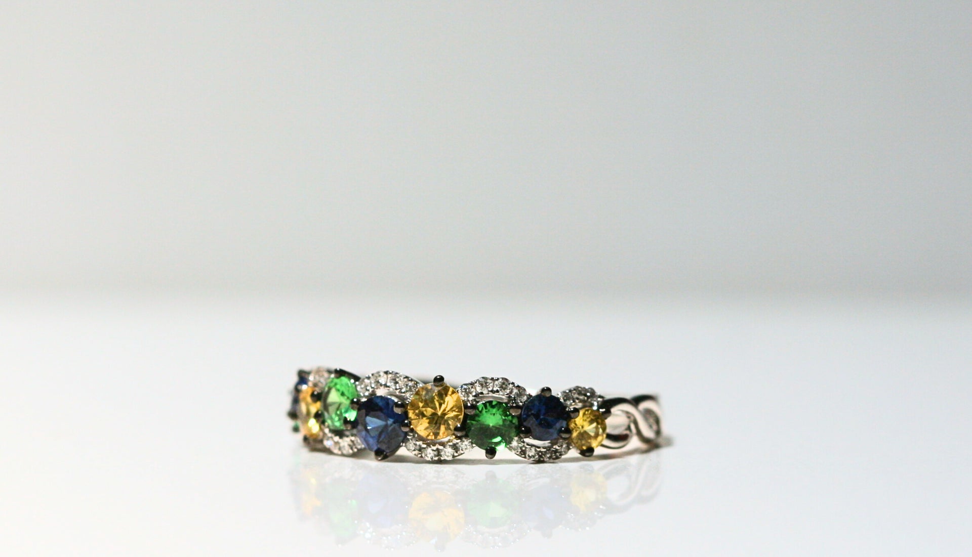 Alternating blue, green, and yellow round stones are framed by curves of white diamonds. All set in white gold - and the prongs are plated in black rhodium. The sides of the band are twisting strands of white gold.