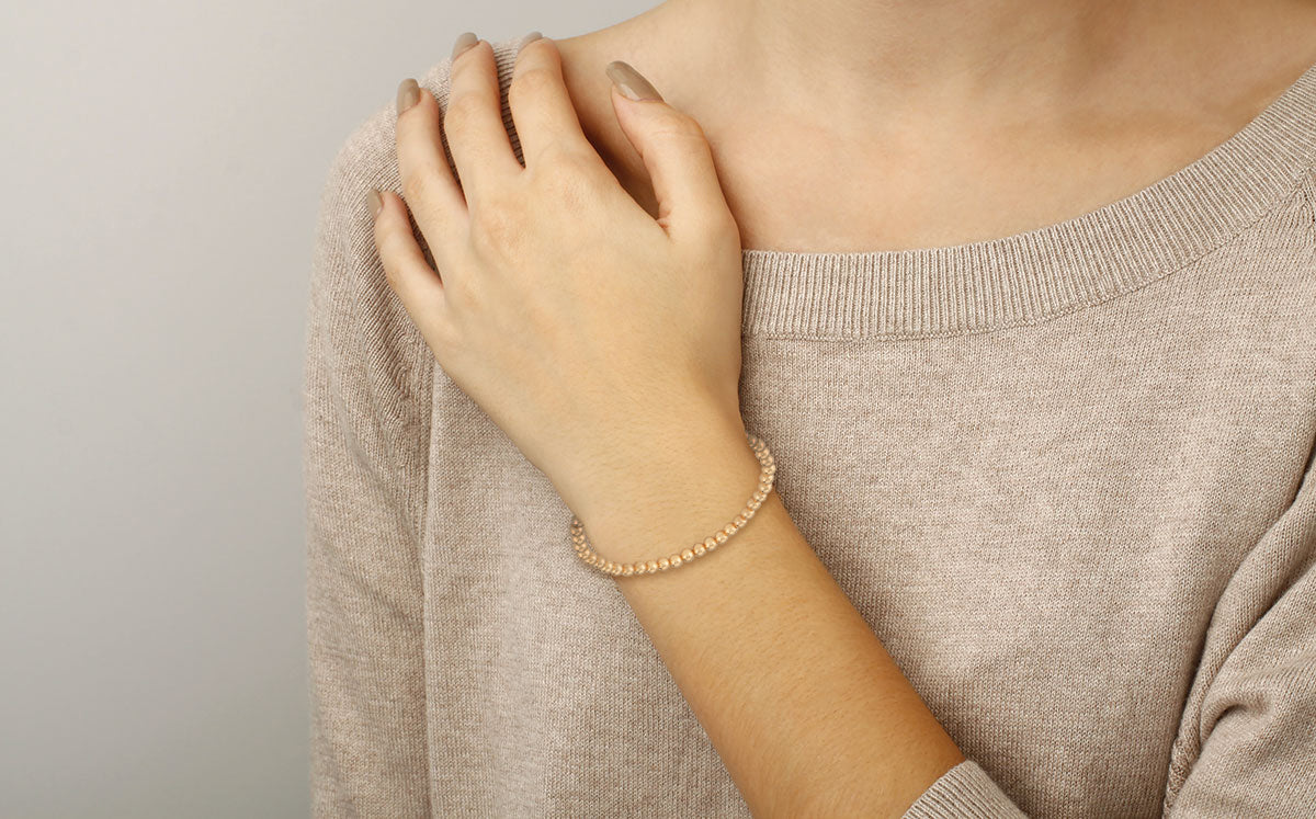 Person wearing a beige sweater and gold beaded bracelet on a plain background