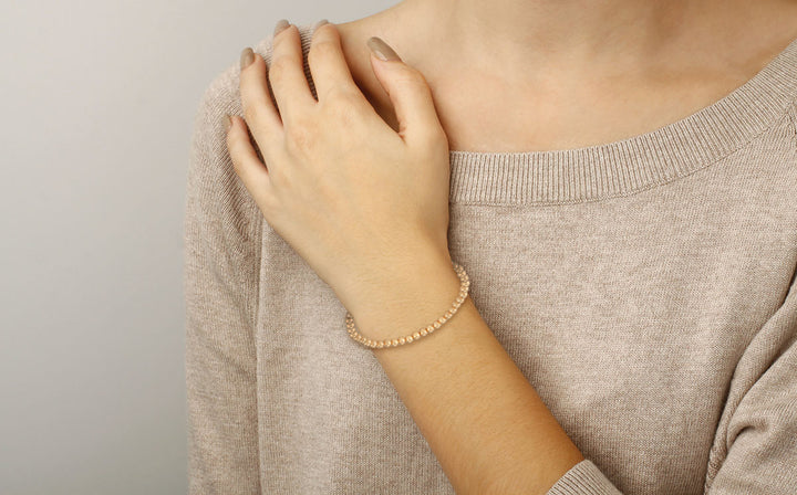 Person wearing a beige sweater and gold beaded bracelet on a plain background