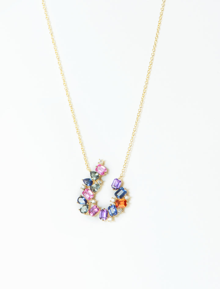 Pendant of multicolored gemstones hangs off a yellow gold chain. The stones are different shapes - emerald cut, pear cut, and round diamonds. The stones are blue, green, orange, pink, and purple. The stones are arranged in an open circle.