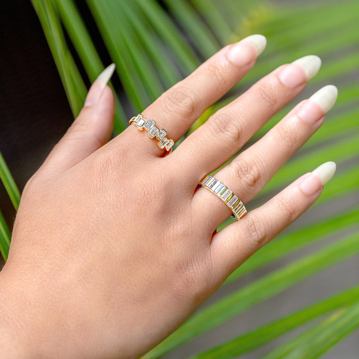 A close-up of a hand wearing two gold diamond rings. One ring is staggered emerald cut diamonds while the second one is a straight band of channel set straight baguette diamonds, set against a background of green palm leaves.