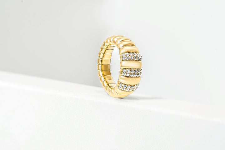 A yellow gold ring with a textured, segmented design and rows of clear pavé diamonds, standing upright on a white surface against a light gray background.