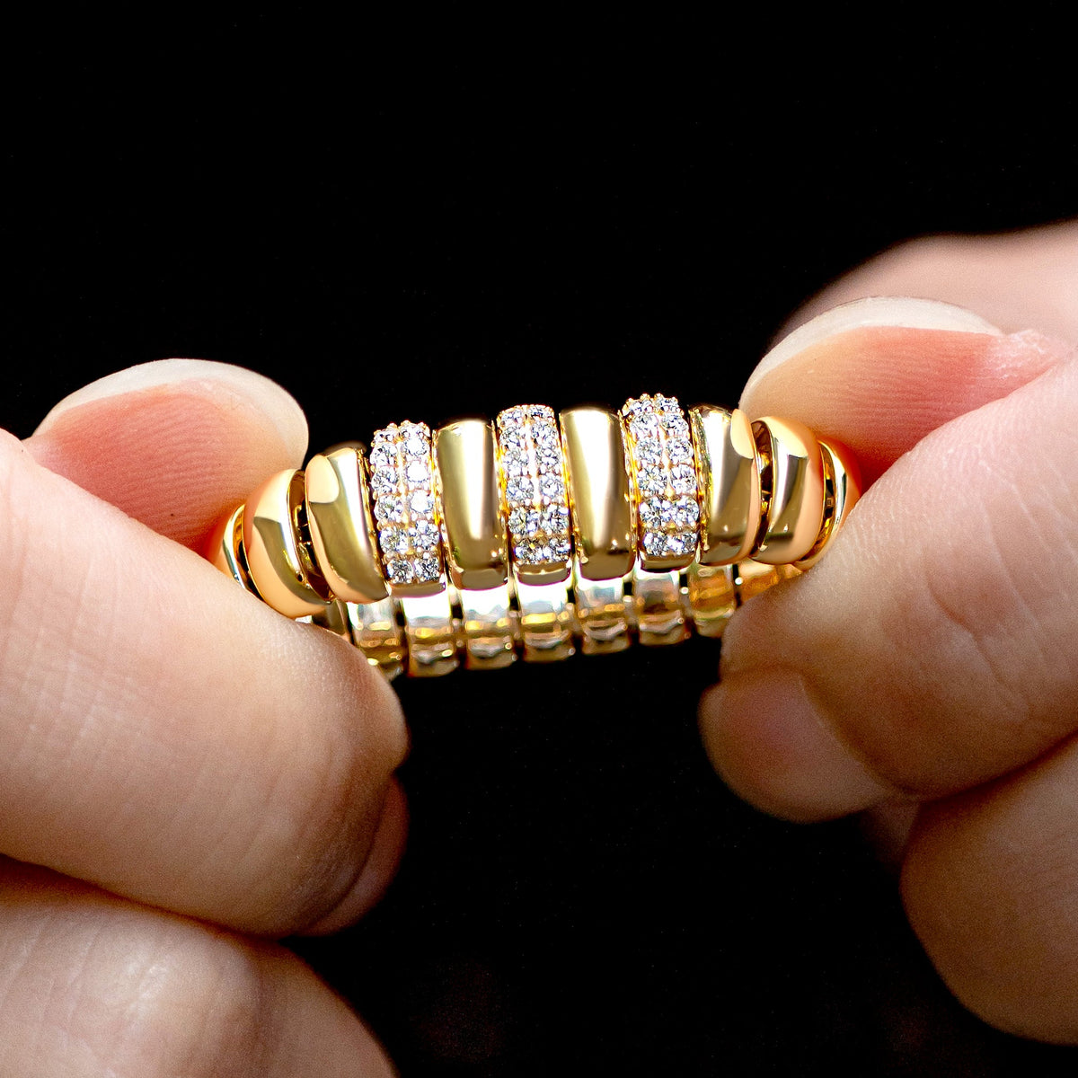 A close-up of a gold flexible stretch ring with multiple rows of pavé-set clear stones, held between two hands against a solid black background.
