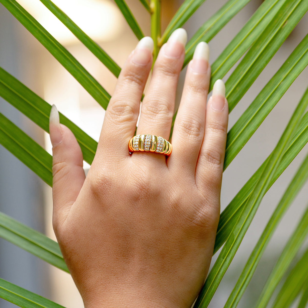 A gold ring with three vertical rows of small pave diamonds worn on a person's ring finger, shown against a background of green palm leaves.