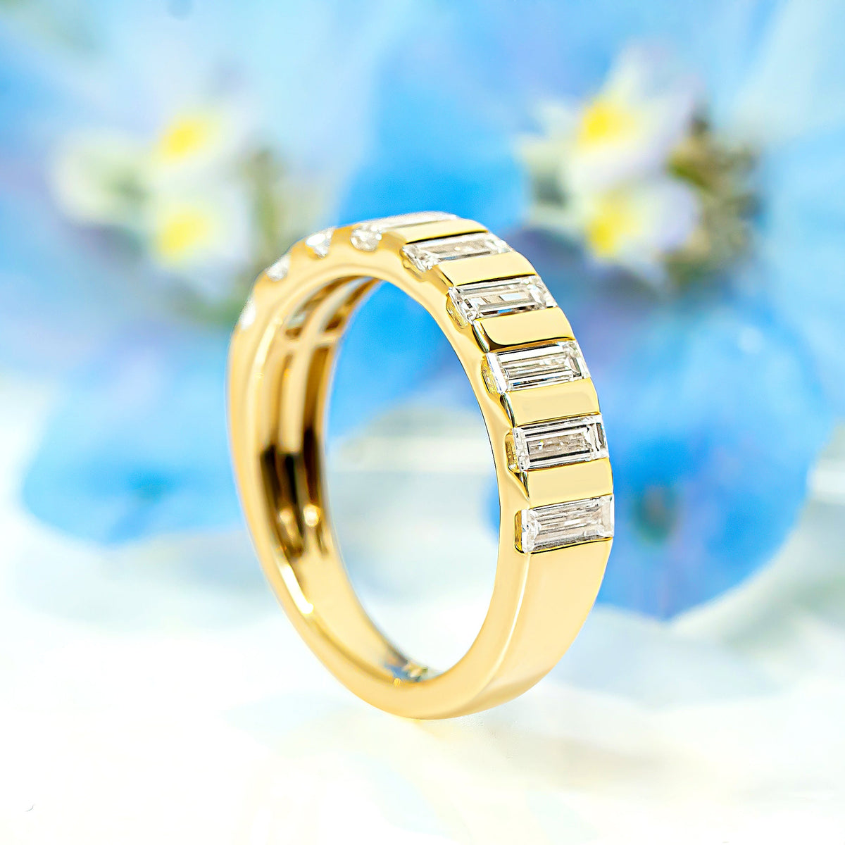 A yellow gold ring featuring baguette-cut diamonds, standing upright against a soft-focus background of blue flowers.