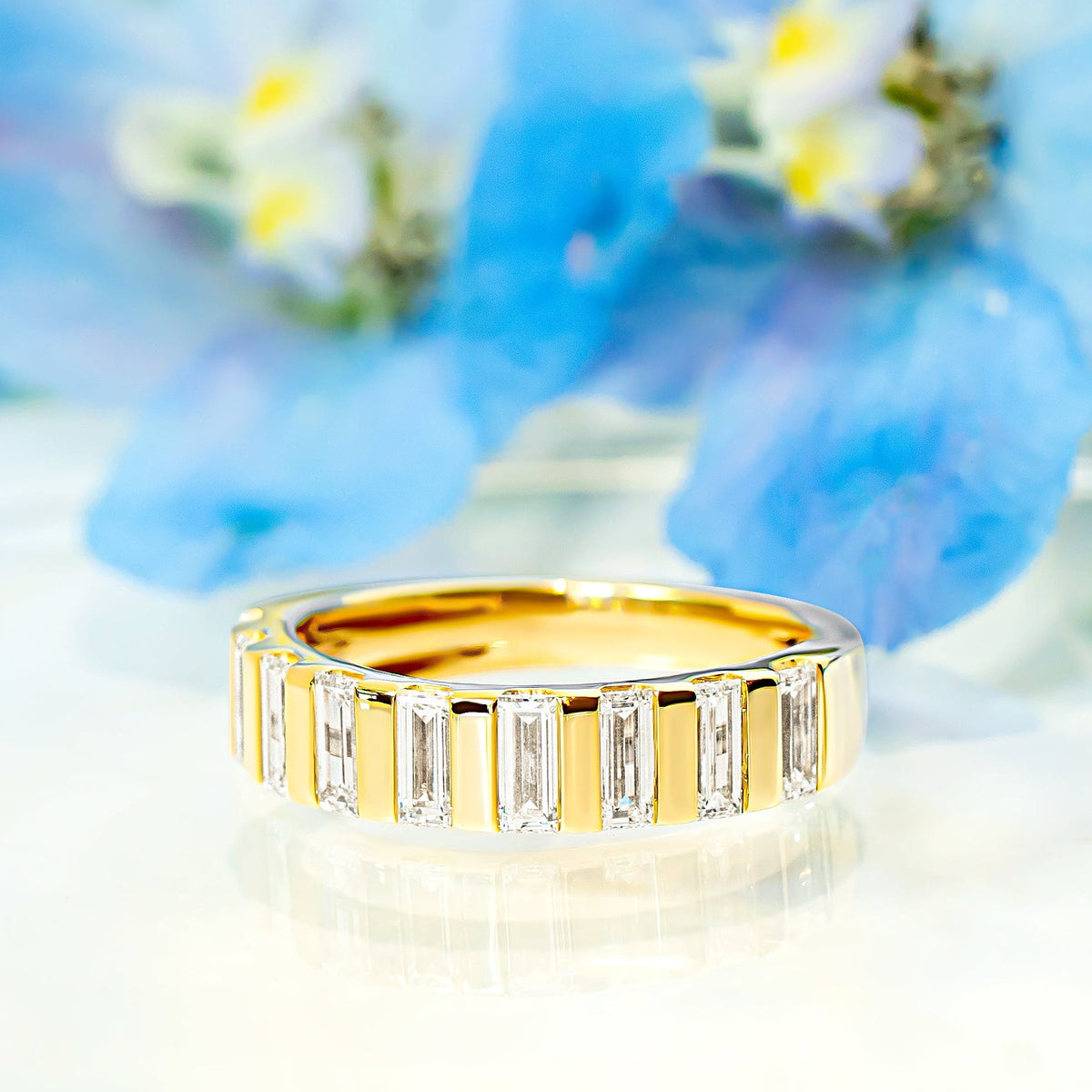 A gold band ring featuring a row of baguette-cut diamonds, photographed on a glossy white surface with soft-focus blue flowers in the background.