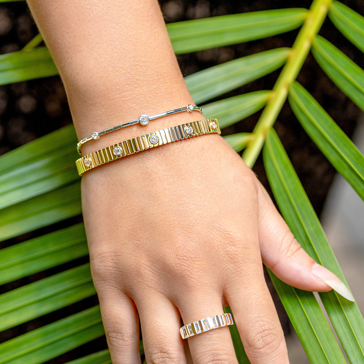 A close-up of a hand wearing a gold channel-set baguette diamond band and a gold textured diamond bangle bracelet, with a thin white gold diamond station bracelet, styled against green palm leaves.