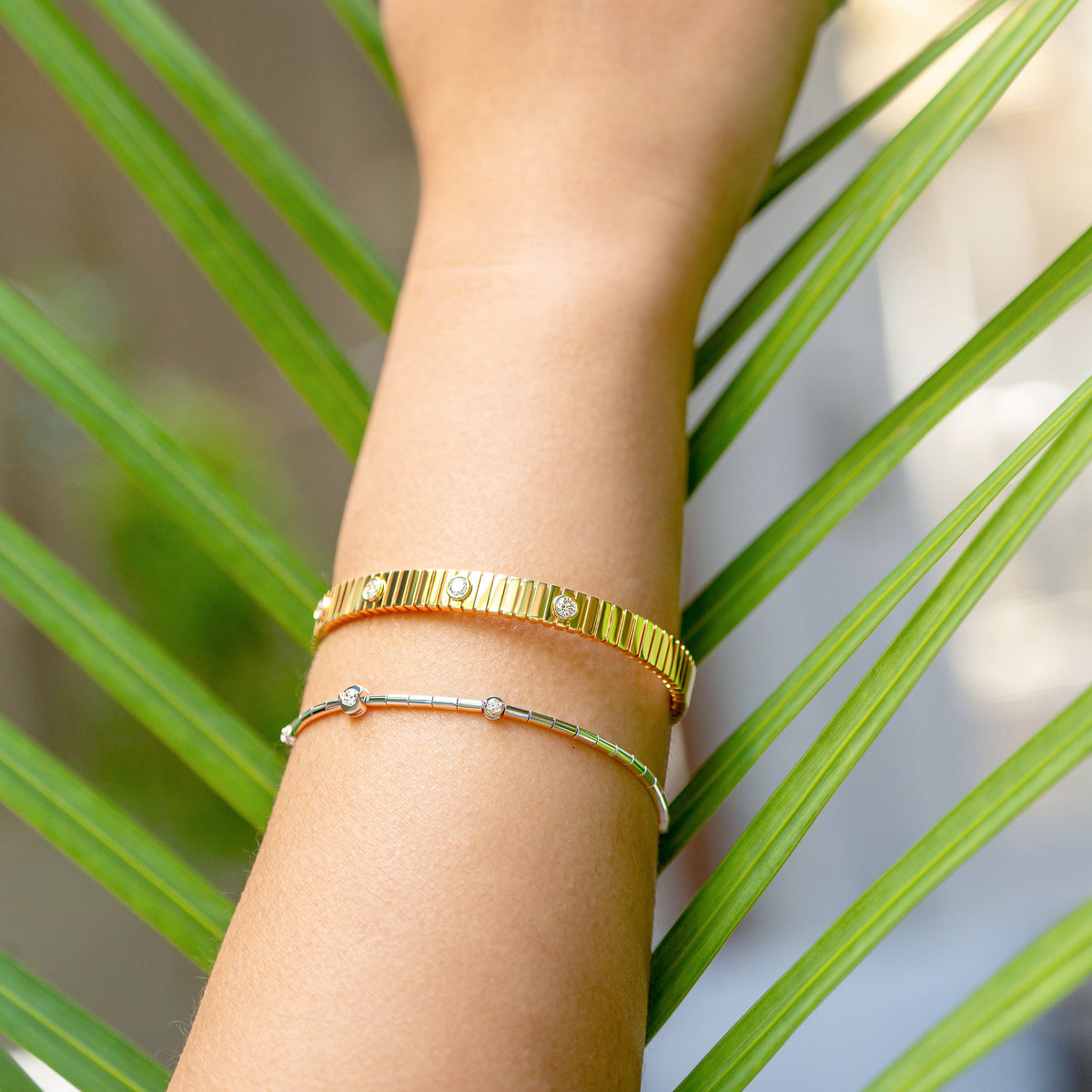 A close-up of a person's hand and wrist adorned with gold jewelry, including a gold fluted diamond bangle and a white gold diamond station line bracelet, set against a background of green palm leaves.