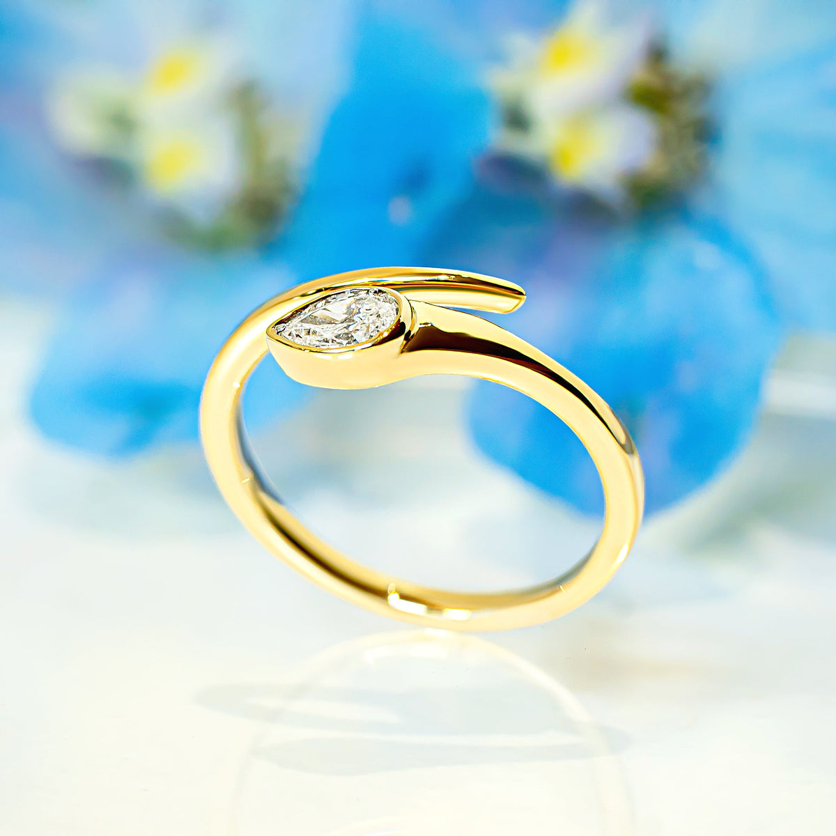 A gold ring with a pear-shaped diamond, displayed on a reflective white surface with a soft-focus background of blue flowers.