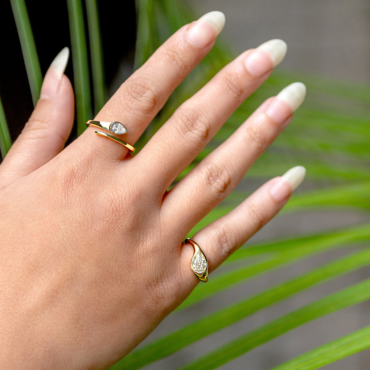 A close-up of a hand wearing two gold diamond rings. One open style ring with a pear shape diamond and the second ring with an asymmetrically oriented pear cut worn on the pinky, posed in front of green palm leaves. 