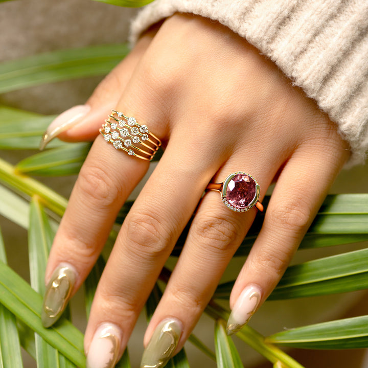 A close-up of a hand wearing two gold rings, one with a rows of round brilliant diamonds and the other with a round berry colored pink zircon, resting on green palm leaves with a beige knit sleeve visible.