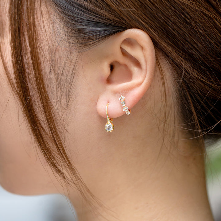 A close-up side profile of a woman's ear wearing a delicate gold huggie hoop earring with a bezel-set round brilliant cut drop down and a huggie hoop of various cut diamonds on her lobe.