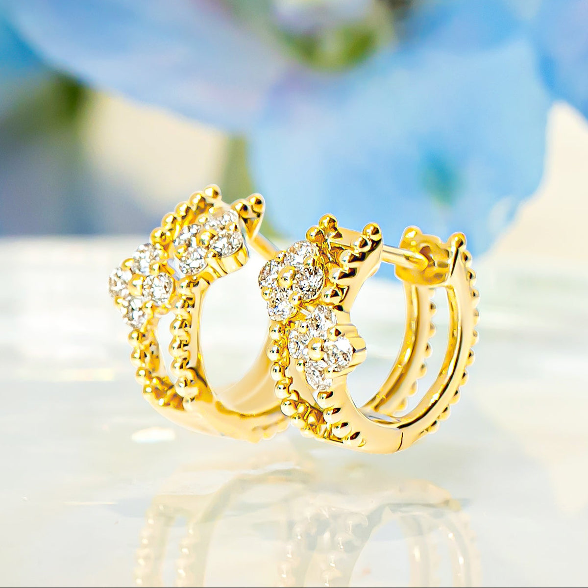 A pair of gold huggie hoop earrings adorned with diamond floral accents, displayed on a reflective white surface with a soft-focus blue floral background.