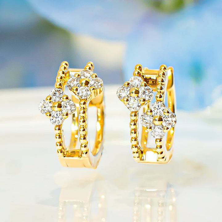 A pair of gold huggie hoop earrings with diamond floral accents, displayed on a reflective white surface with a soft-focus blue background.