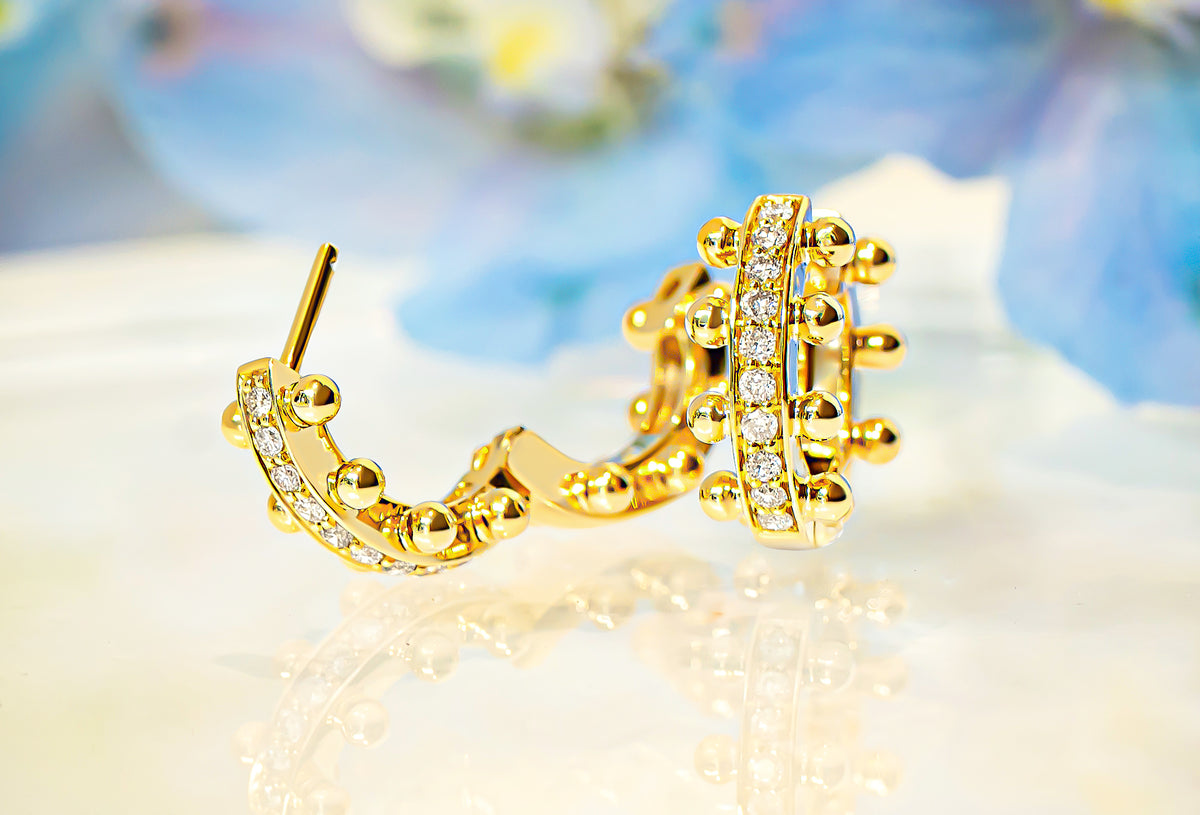 A pair of gold hoop earrings with diamonds and gold beaded detailing, displayed on a reflective white surface with a soft, blurred blue and white background.
