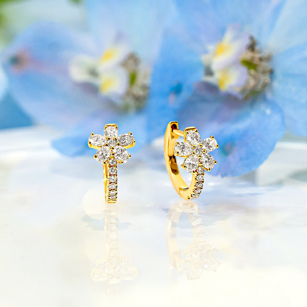 A pair of gold hoop earrings with small diamonds in a flower shape, displayed on a clear glass plate with blue flowers in the background.