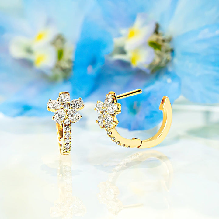 A pair of gold hoop earrings with floral diamond designs, displayed on a reflective white surface with soft-focus blue flowers in the background.
