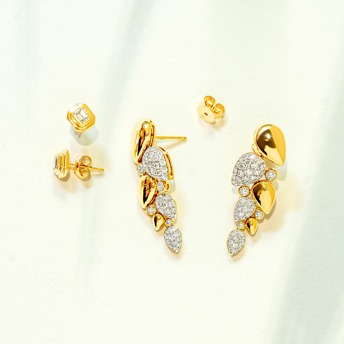 Gold and diamond leaf-shaped drop earrings and bezel emerald-cut diamond stud earrings displayed on a soft white background with subtle green leaf shadows.