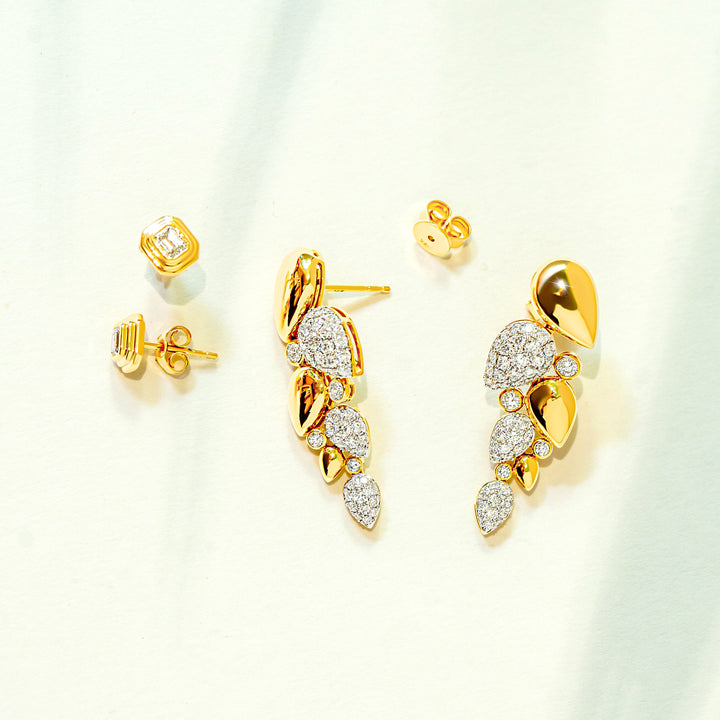 Gold and diamond leaf-shaped drop earrings and bezel emerald-cut diamond stud earrings displayed on a soft white background with subtle green leaf shadows.