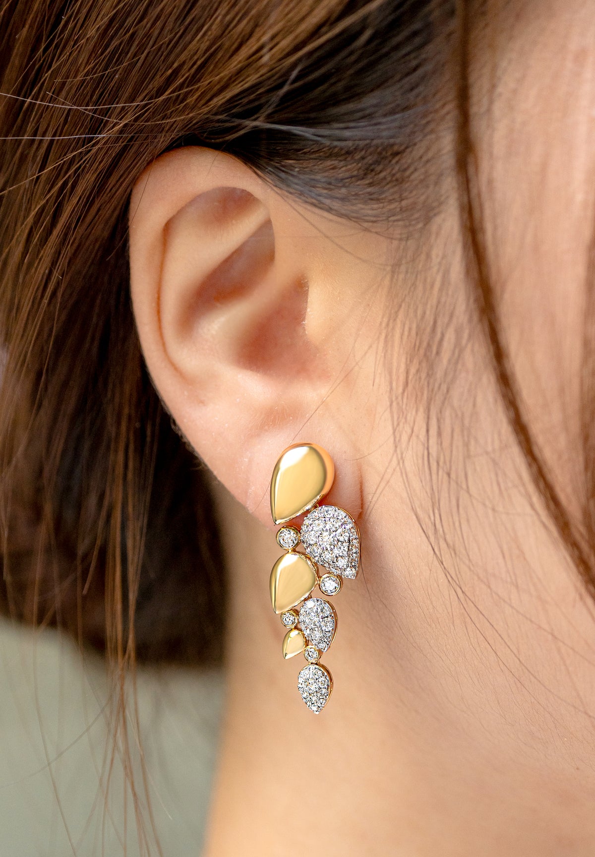 A close-up of a person's ear wearing a gold and white gold drop earring with organic-shaped design and pave diamond accents. 