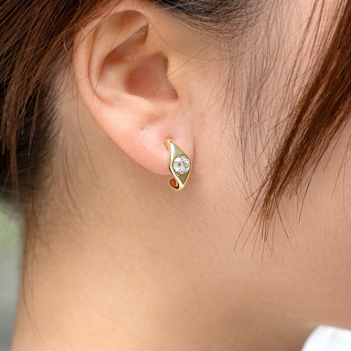 A close-up of a person's ear wearing a small gold crescent-shaped stud earring with a white round diamond. 