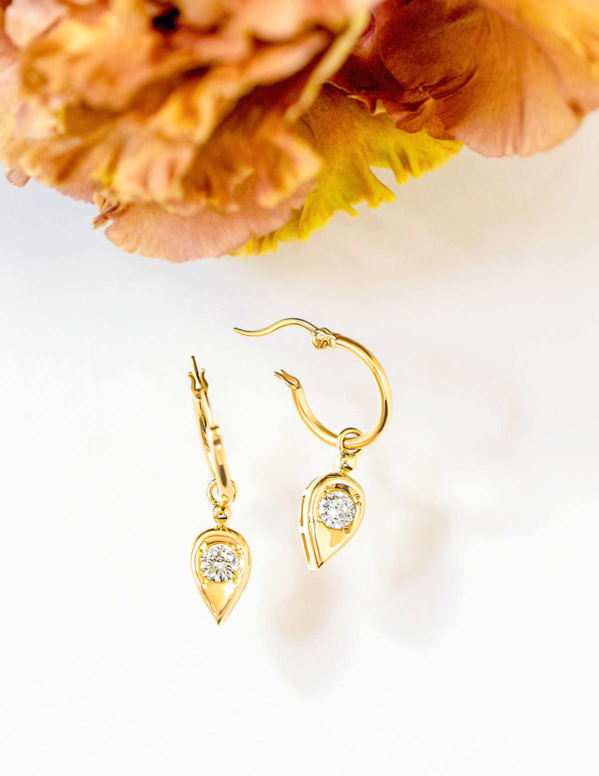 A pair of gold drop hoop earrings with teardrop-shaped dangles set with round brilliant cut diamonds, displayed on a white surface with a blurred orange flower in the background.
