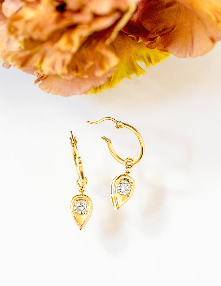 A pair of gold drop hoop earrings with teardrop-shaped dangles set with round brilliant cut diamonds, displayed on a white surface with a blurred orange flower in the background.