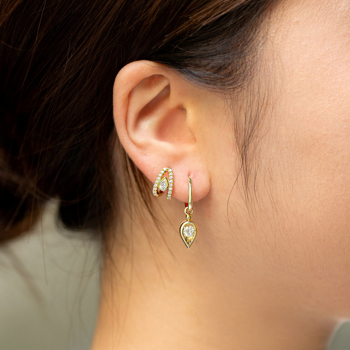 A close-up side profile of a woman wearing a pair of small gold hoop earrings with a pear diamond and a dangling gold teardrop earring with a round diamond, against a soft blurred background.