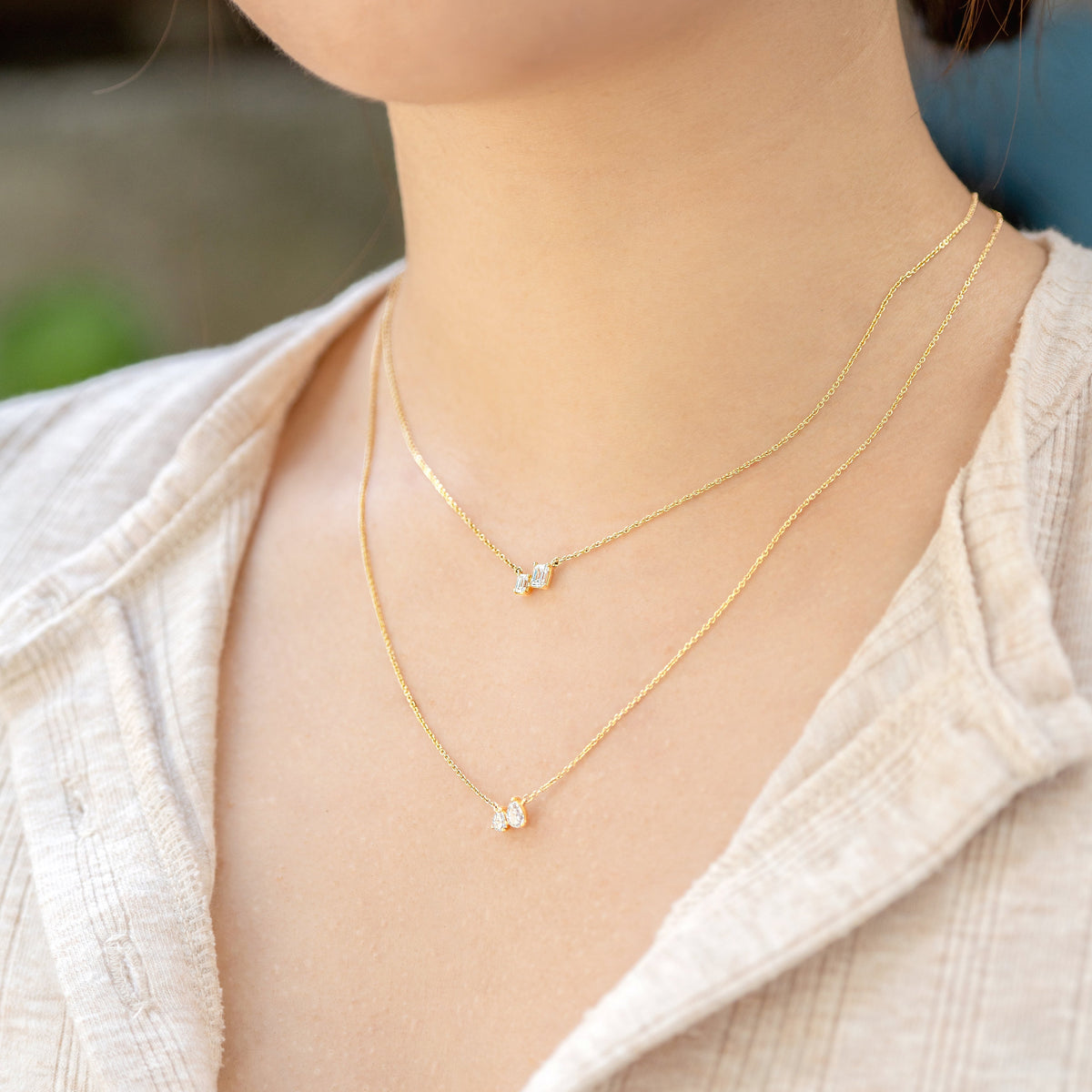A close-up of a person wearing two delicate gold necklaces with moi et toi style diamonds, styled with a light beige linen shirt.