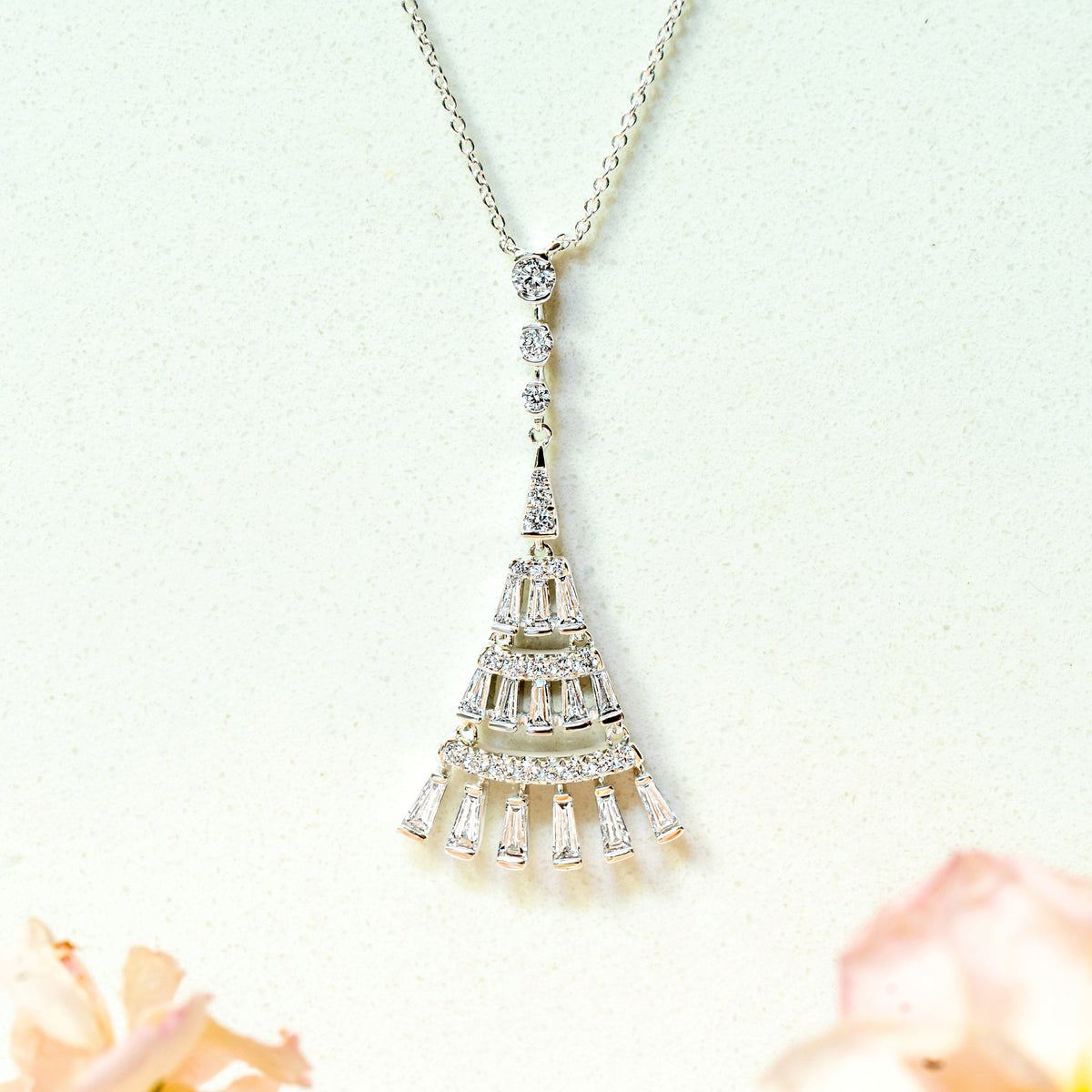 A white gold pendant necklace with a tiered, art-deco design and dangling baguette diamonds, suspended against a light background with soft pink flowers blurred at the bottom.