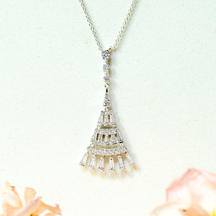 A white gold pendant necklace with a tiered, art-deco design and dangling baguette diamonds, suspended against a light background with soft pink flowers blurred at the bottom.