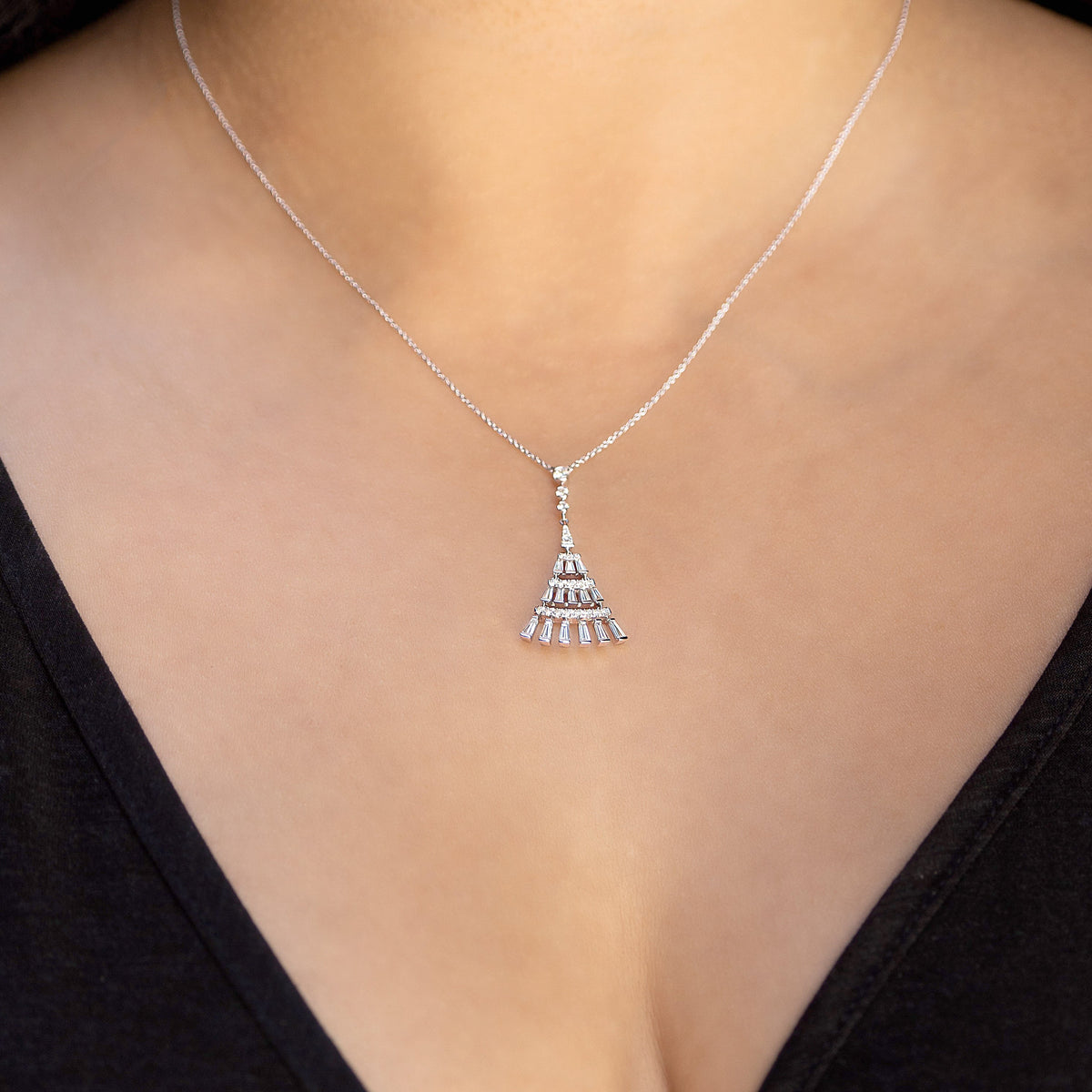 A close-up of a model's neck and chest wearing a delicate silver chain necklace with a small silver-tone tiered, fan-shaped pendant featuring dangling white diamonds stones, paired with a black V-neck top.