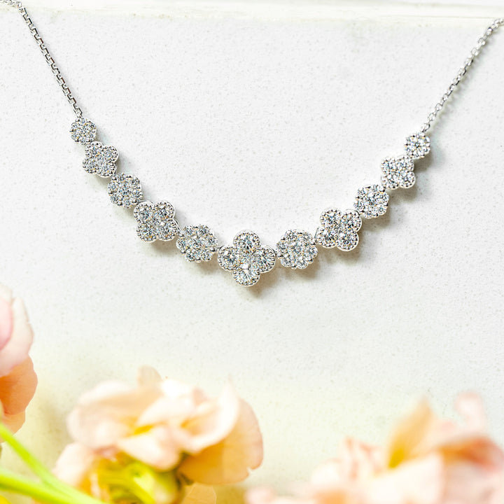 A white gold necklace with a row of sparkling diamonds in clover motifs, displayed on a white surface with soft pink flowers in the foreground.