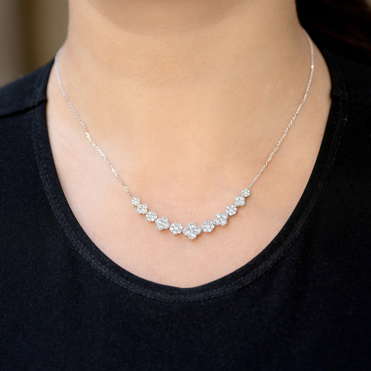 A close-up of a woman wearing a delicate white gold necklace with a curved row of diamond clusters and diamond clover motifs across the chest, paired with a black top.