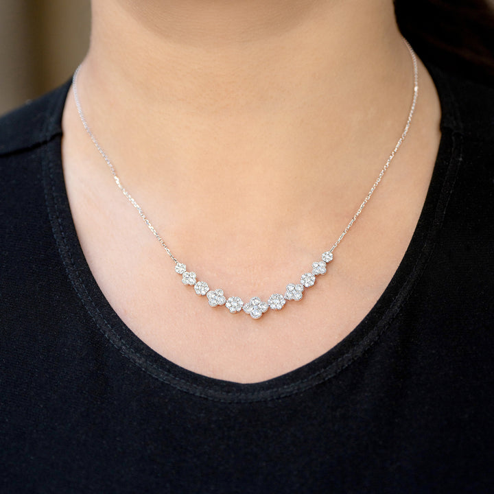 A close-up of a woman wearing a delicate white gold necklace with a curved row of diamond clusters and diamond clover motifs across the chest, paired with a black top.