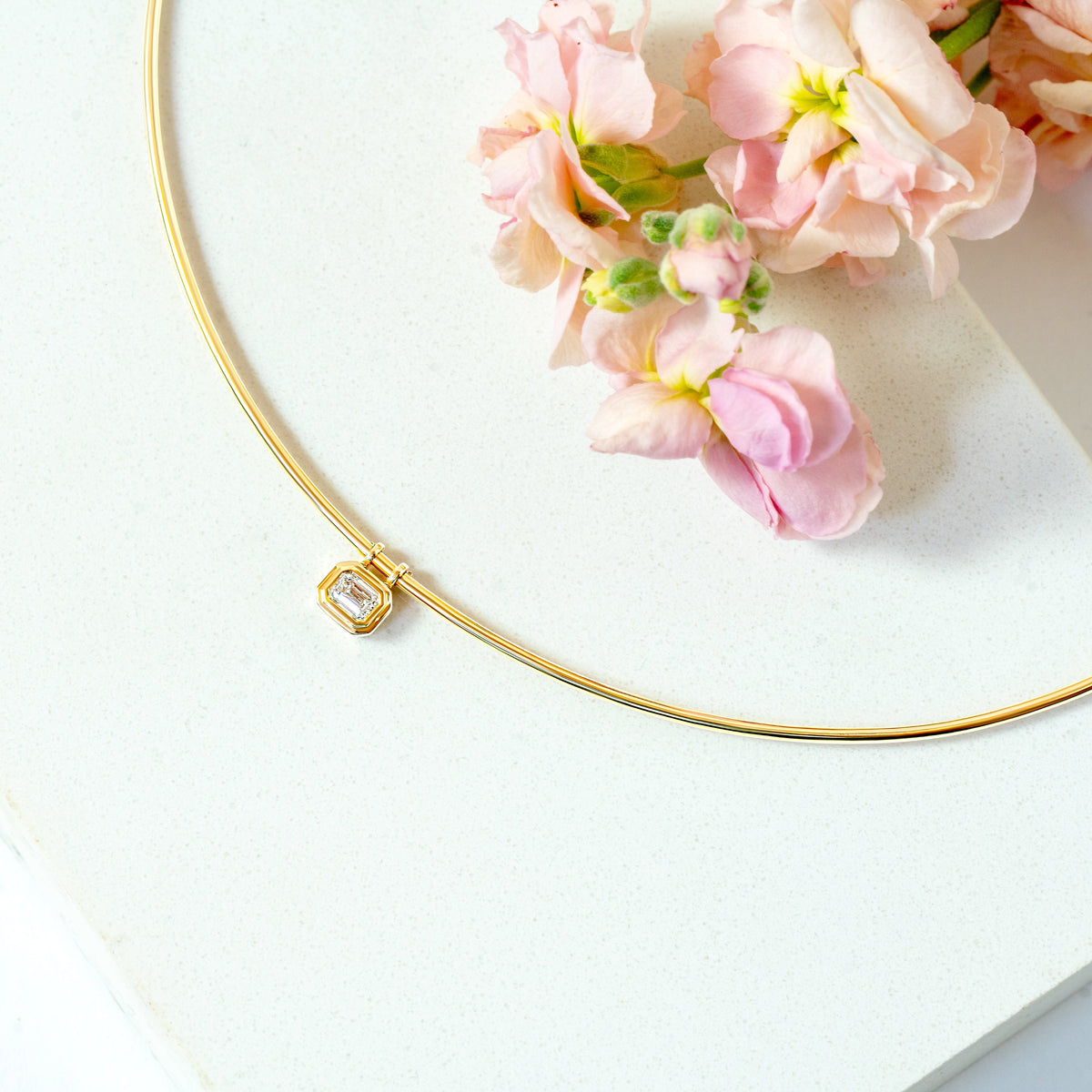 A yellow gold wire necklace with a small emerald-cut bezel-set diamond, styled on a white textured surface next to pink flowers.