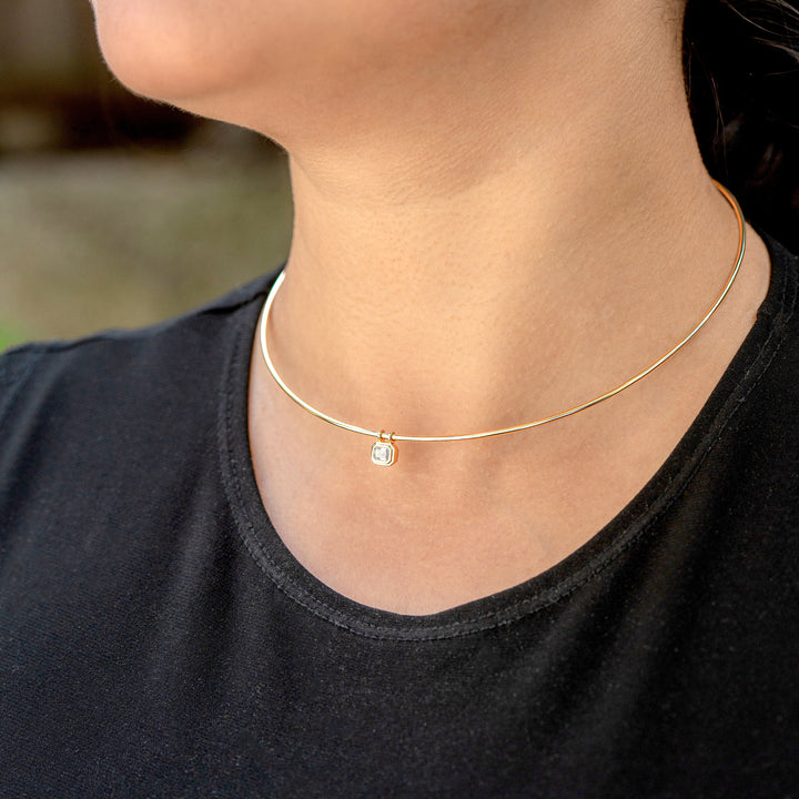 A close-up of a person's neck and lower face wearing a delicate gold wire omega necklace with a bezel set emerald cut diamond pendant, paired with a black t-shirt.