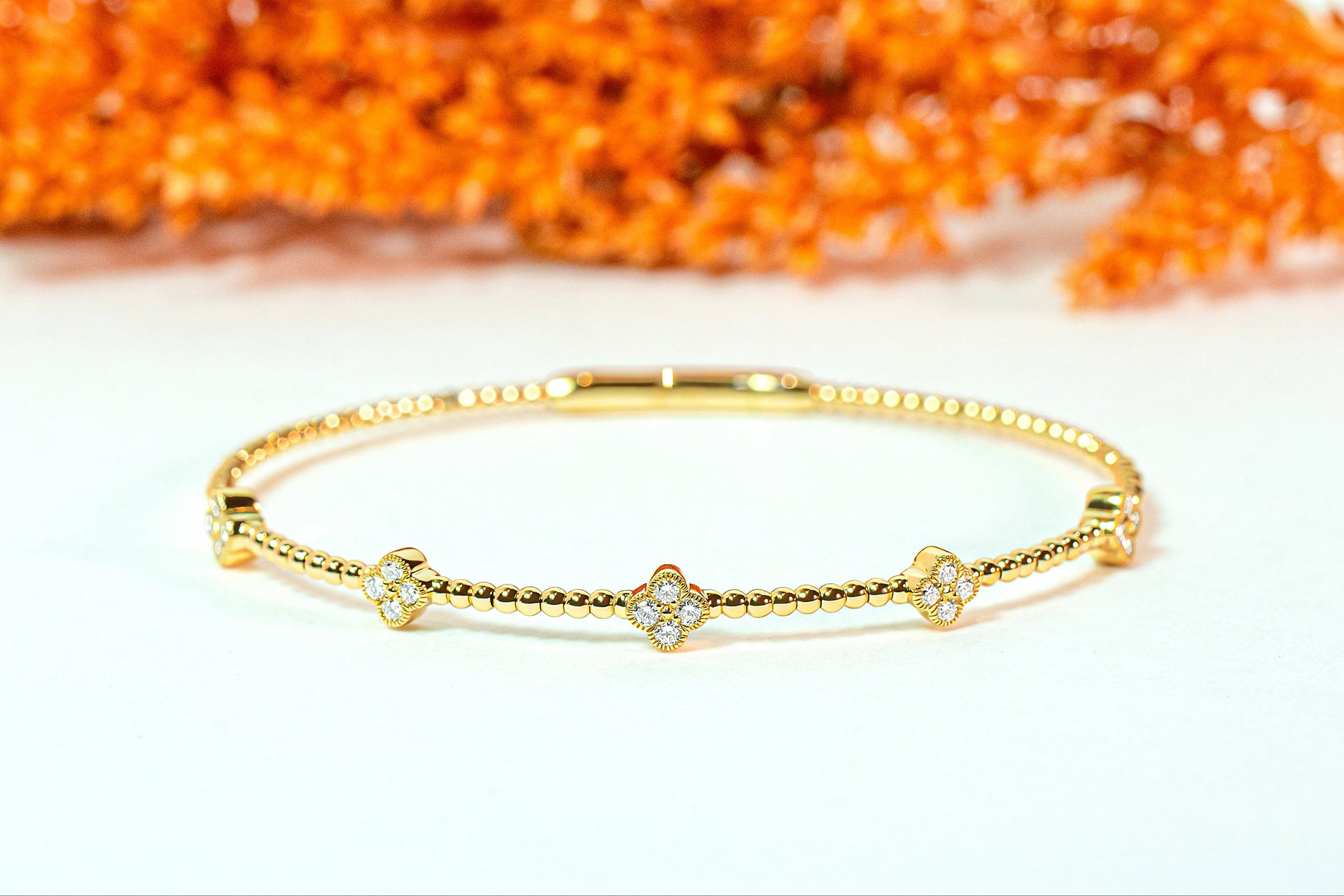 Gold bracelet with diamond clover-shaped charms on a white surface with orange leaves in the background