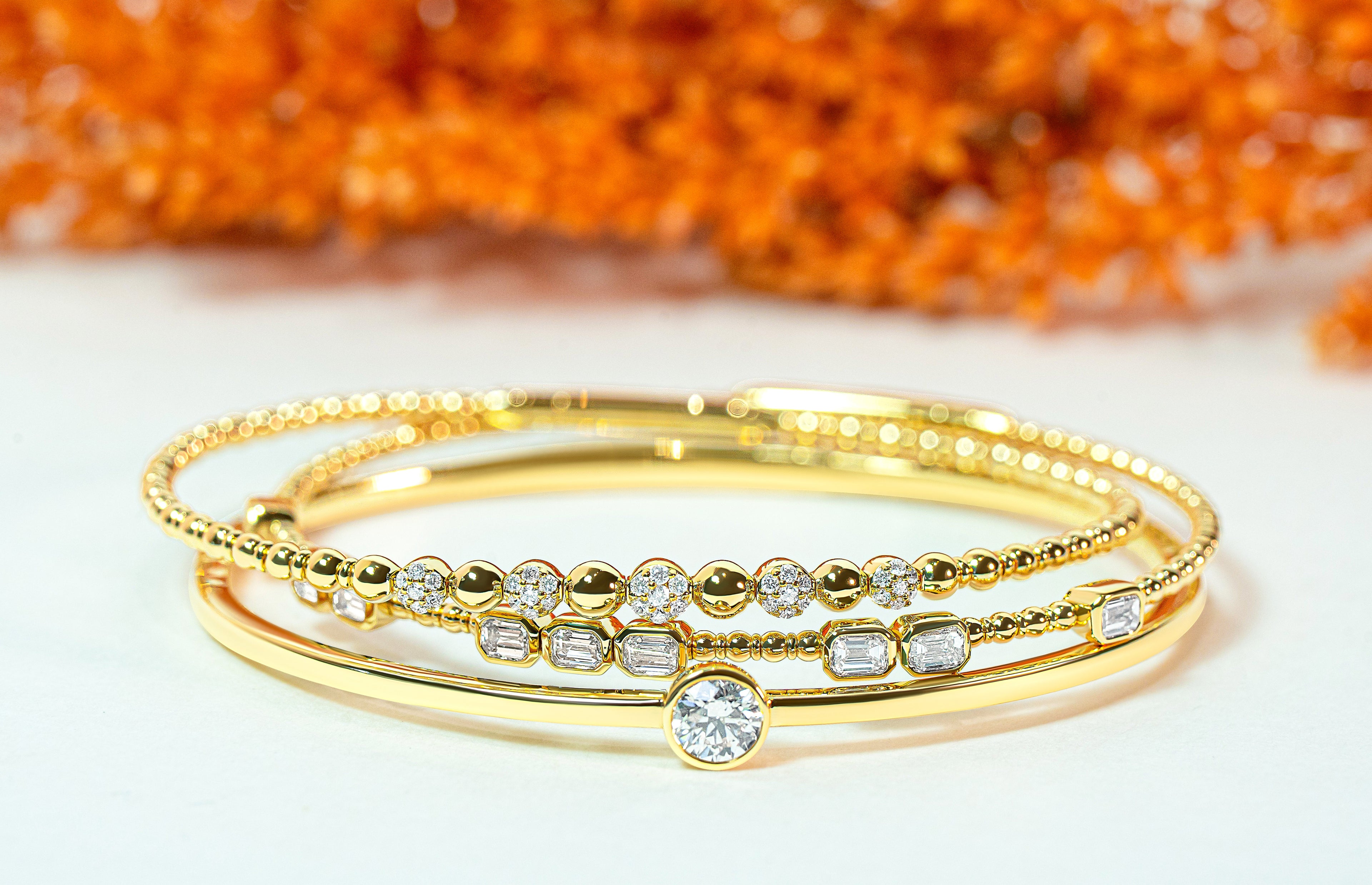 Set of three gold bangles with diamond accents on a white surface with orange leaves in the background.