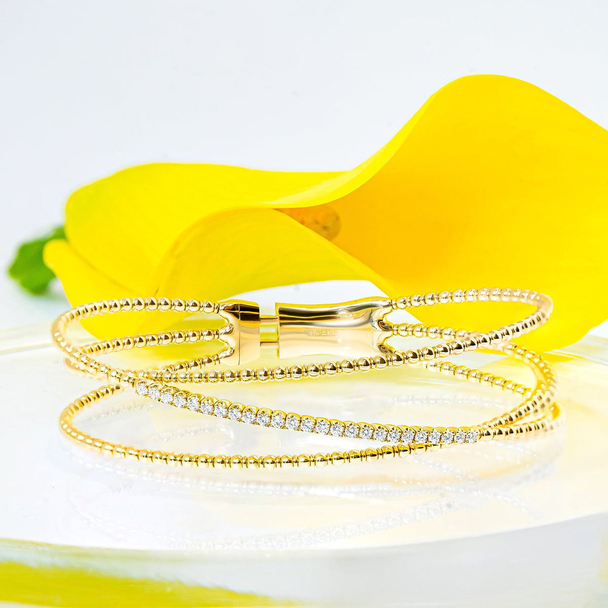 A gold bangle bracelet with three strands, including a row of diamonds, displayed on a clear glass dish with a bright yellow flower behind them against a white background.
