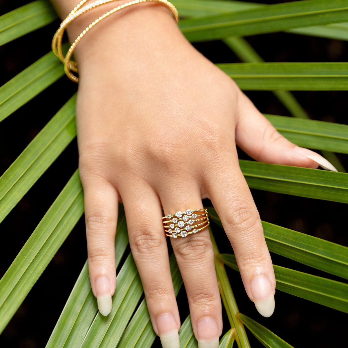 A close-up of a hand resting on green palm leaves, wearing a multi-band gold ring with rows of round brilliant cut diamonds and matching gold bangles.