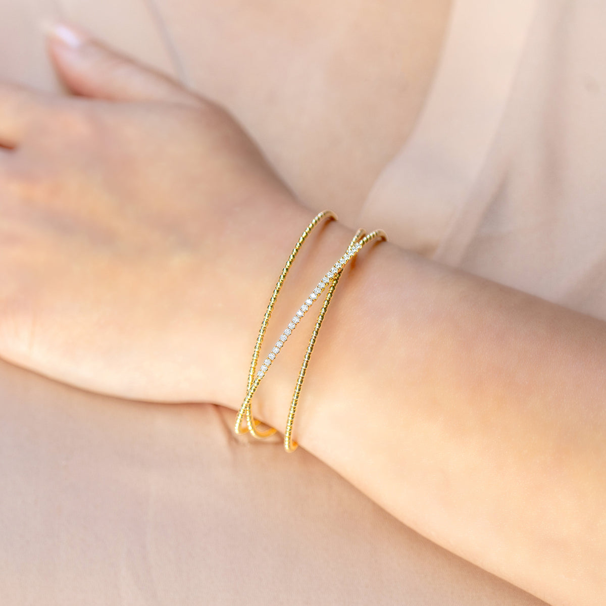A close-up of a person's wrist wearing two delicate bracelets: one gold-toned textured bangle and one thin row of diamonds, set against a soft beige background.