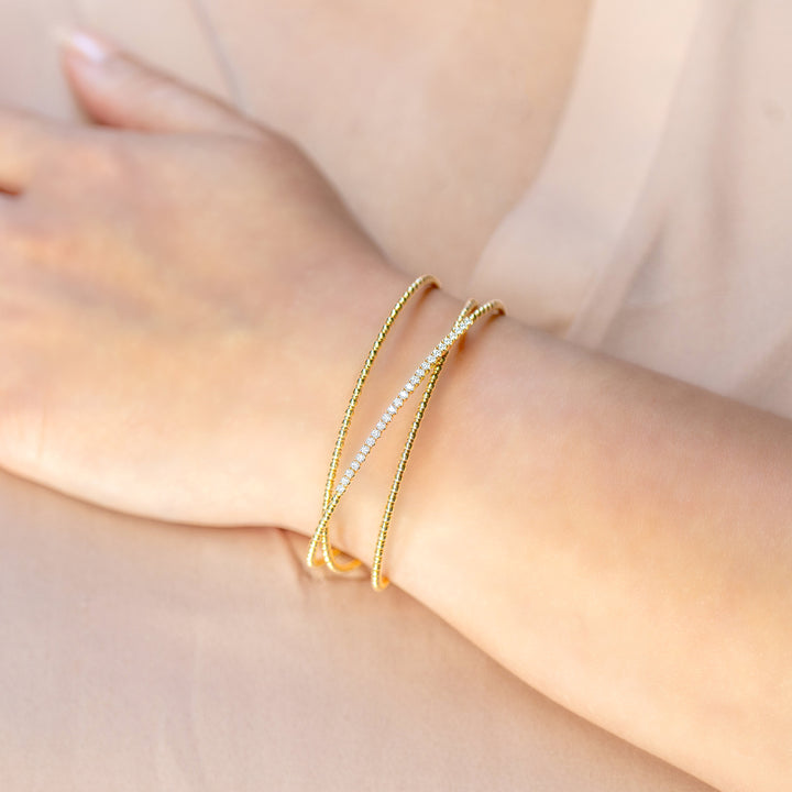 A close-up of a person's wrist wearing two delicate bracelets: one gold-toned textured bangle and one thin row of diamonds, set against a soft beige background.