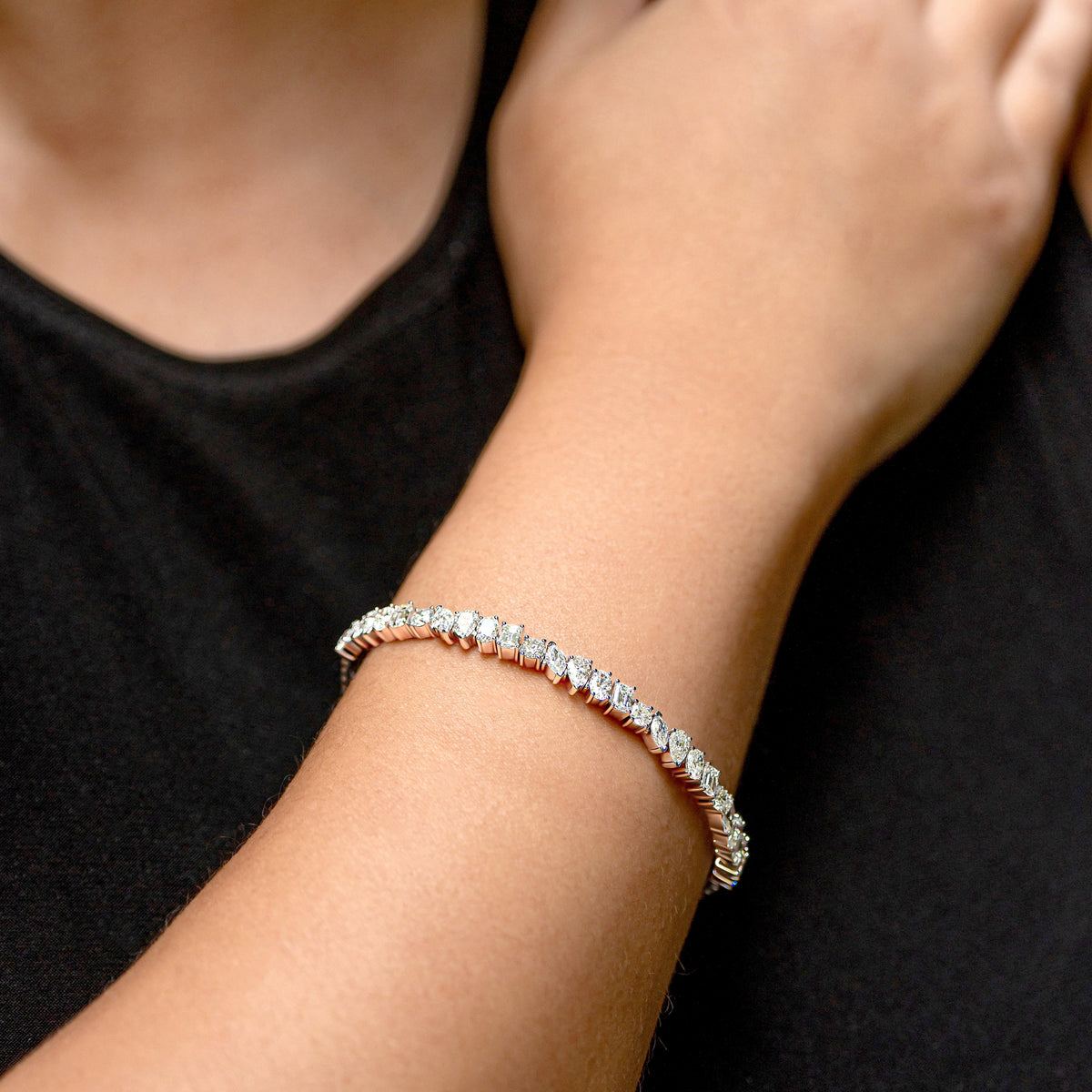 A close-up of a person's wrist wearing a thin silver tennis bracelet with sparkling mixed-cut diamonds, set against a black shirt background.